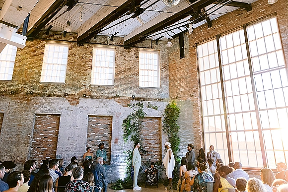 Wedding ceremony with couple exchanging vows beneath a greenery installation, guests seated in a brick-walled loft with large grid windows.