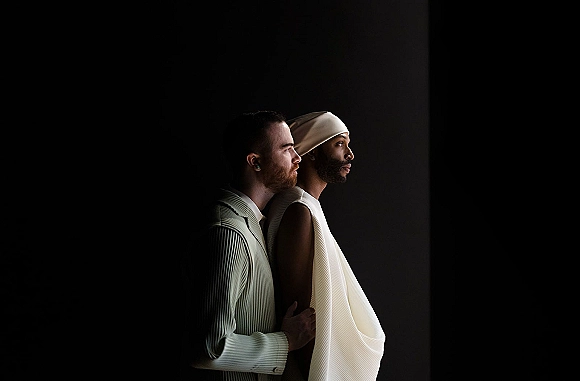 Couple portrait of two grooms standing close in side profile, one in a head wrap and earrings, against a dark studio backdrop