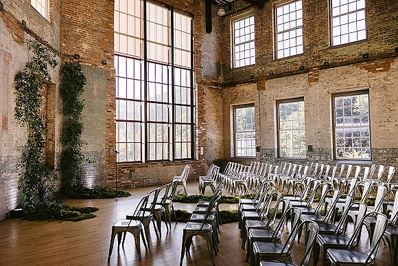 Ceremony setup with metal chairs and a greenery garland aisle in an industrial loft, framed by exposed brick walls and tall windows.