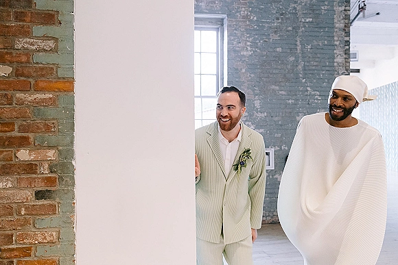 Groom portraits of two same sex grooms indoors, one in a striped suit with boutonniere, posed by an industrial brick wall and window light