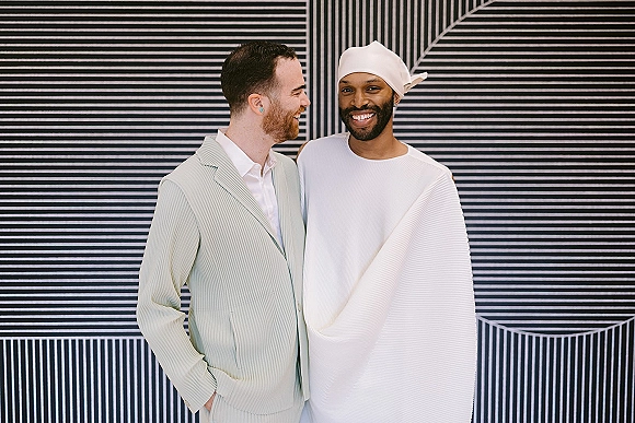 Couple portrait of two grooms smiling together, one in a light suit and the other in a white head wrap, against a striped wall backdrop