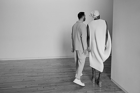 Couple portrait holding hands, bride in wedding dress with headwrap and platform shoes, groom in suit, in a minimalist empty room