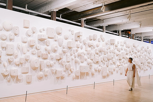 Groom portrait in a white suit with a sleeveless top and belt, standing on a hardwood floor in an art gallery by a sculptural wall