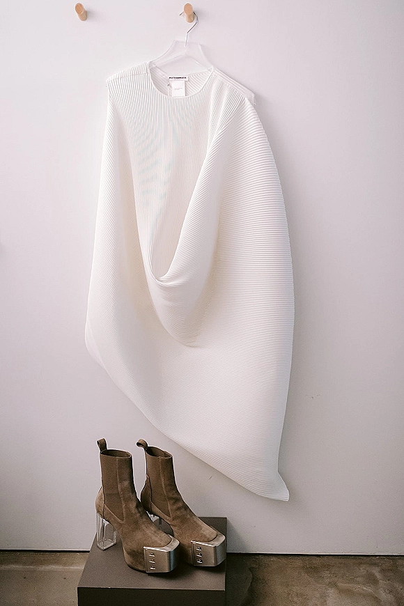 Wedding dress close-up of a modern bridal dress hanging from a wall hook above ankle boots and platform heels against a light gray wall