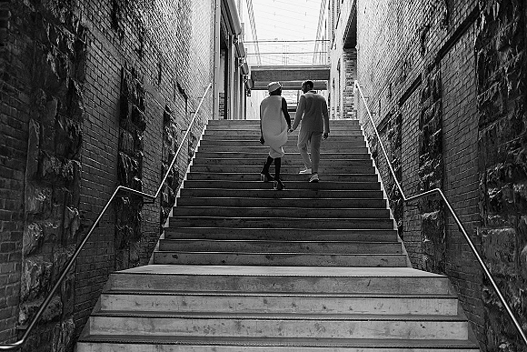 Couple portrait of bride and groom walking away holding hands on concrete stairs, bride in a bridal hat beneath an overhead bridge