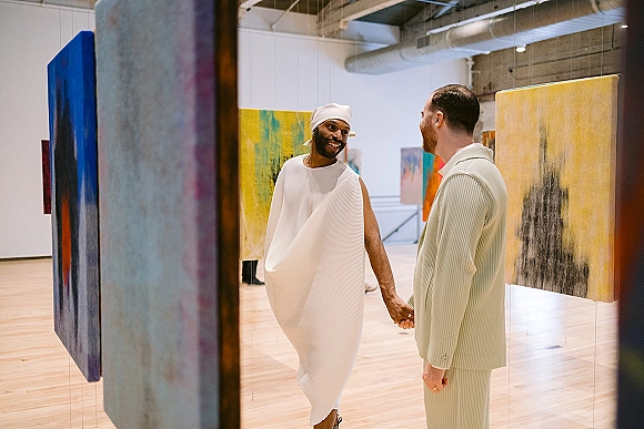 Couple portrait of two grooms holding hands, one in a white headwrap and pleated outfit, the other in a striped suit in an art gallery.