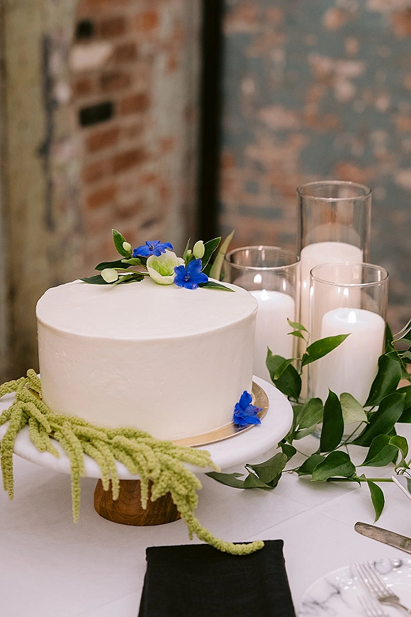Wedding cake with white buttercream frosting on a pedestal stand, topped with blue flowers and greenery, beside candles by a brick wall
