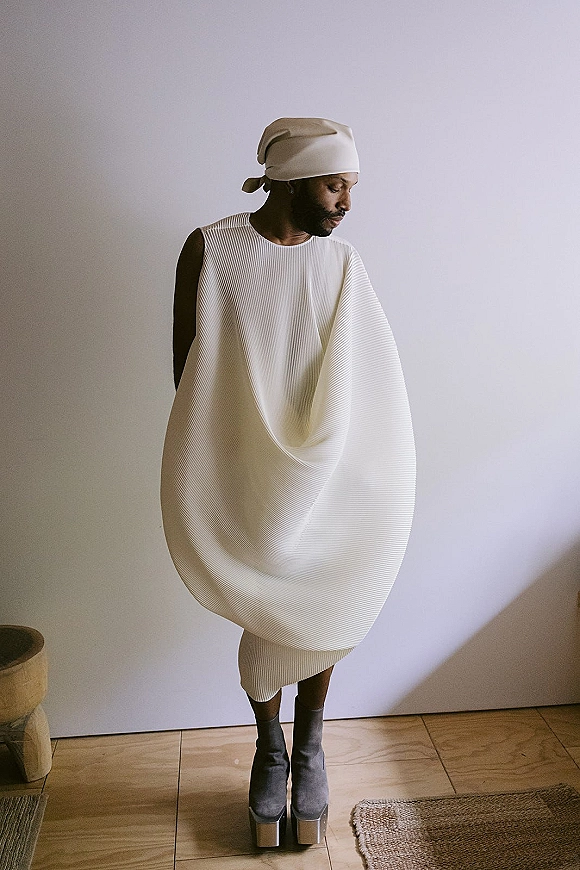 Bridal portrait of an alternative bride look in a white avant garde wedding dress with headwrap and platform boots against a plain wall