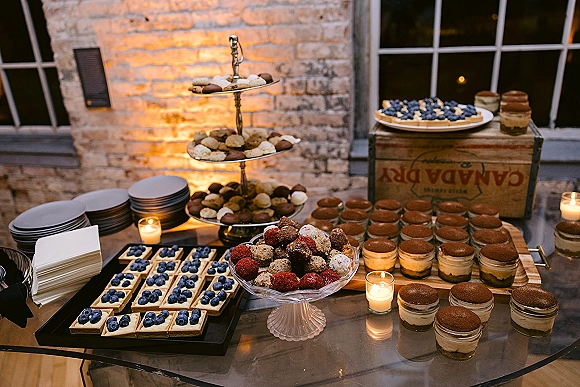 Wedding dessert table with mini dessert cups on tiered stand, fruit tarts and cake truffles, lit by votive candles against brick wall windows