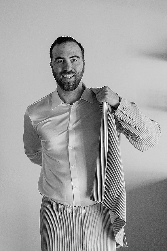 Groom portrait with beard in a dress shirt and striped suit jacket, standing against a plain indoor wall with a relaxed smile