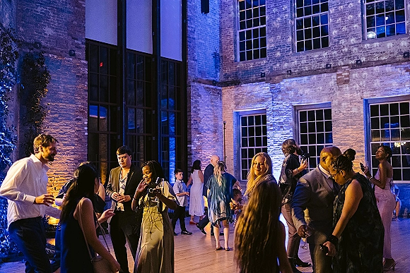 Wedding dance floor packed with reception dancing guests in suits and dresses, holding cocktails under blue uplighting in a brick-walled courtyard venue
