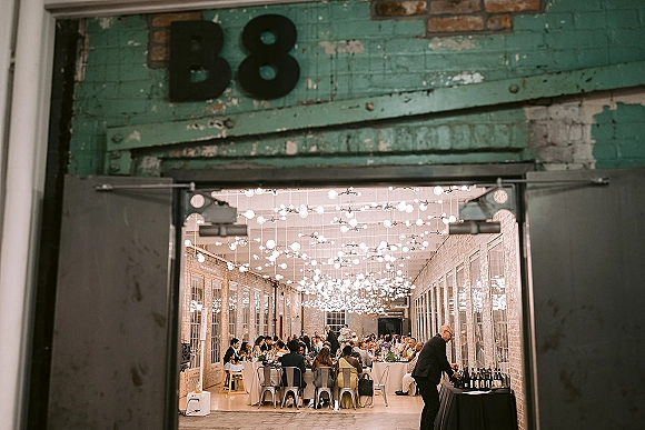 Reception dinner with long banquet tables under string lights, set with glassware and wine bottles in a brick-walled industrial loft