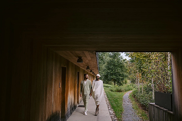 Couple portrait of same sex couple wedding holding hands, one in light green suit, the other in draped dress and headwrap by wood-paneled walkway