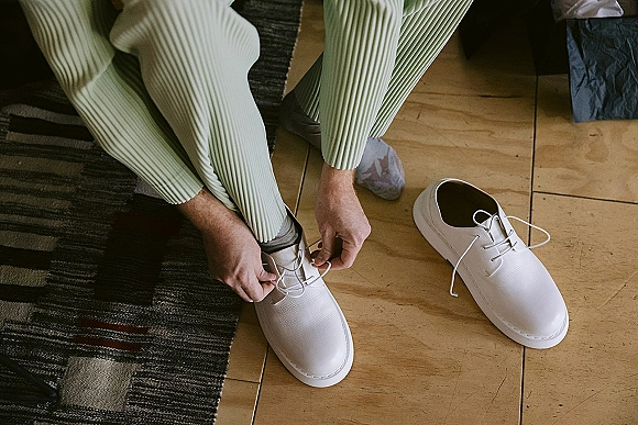 Groom getting ready, tying white dress shoes with patterned socks and pleated pants on a wood floor beside a striped rug