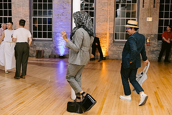 Wedding dance floor reception dance party with guests in suits and sunglasses dancing under uplighting in an industrial loft with brick wall windows