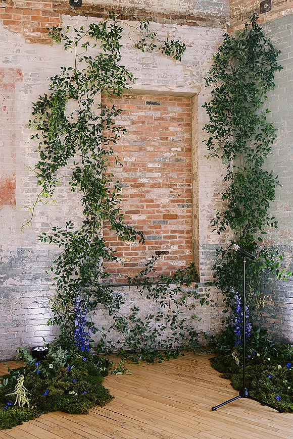 Wedding ceremony backdrop with greenery garland and hanging vines, blue flowers and moss floor decor against an exposed brick wall with uplights