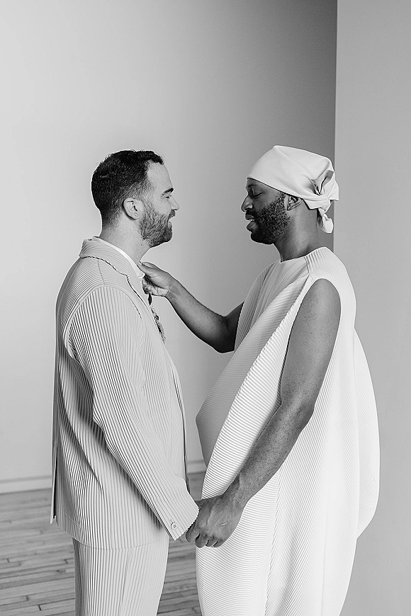 Couple portrait of a same sex wedding couple holding hands as one groom adjusts his tie, in a minimalist studio with wood floor
