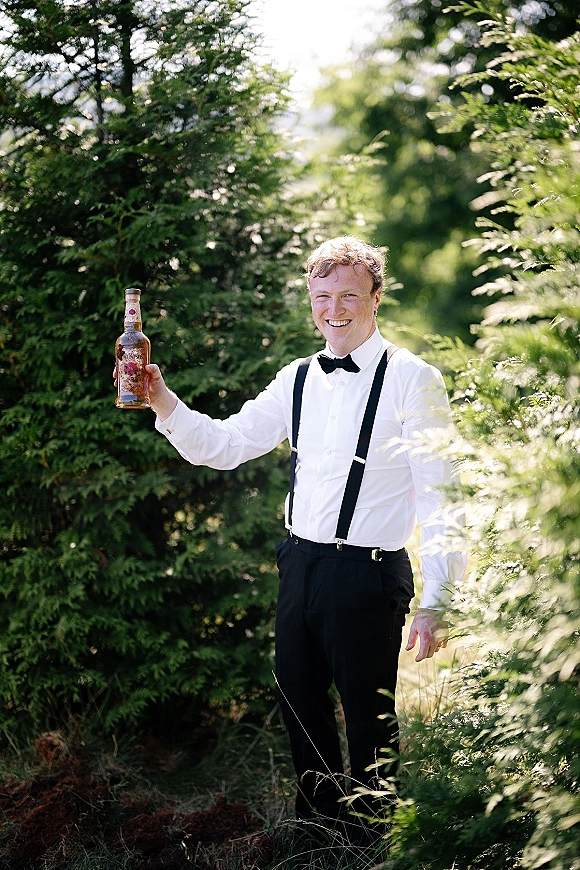 Groom portrait with black bow tie and suspenders, holding a liquor bottle in a white shirt and black trousers amid evergreen forest greenery