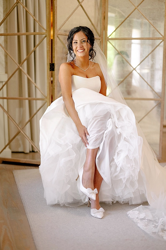 Bridal portrait of a smiling bride in a strapless wedding dress, seated to show bow heels by glass doors with curtains and wood floor