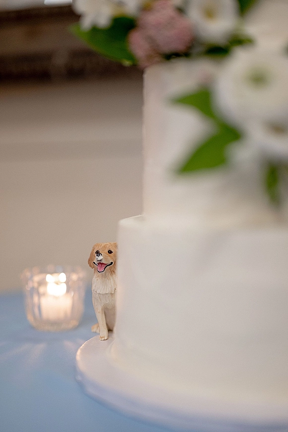Wedding cake with smooth white tiered wedding cake design, floral decoration and dog topper on a light blue tablecloth with votive candle