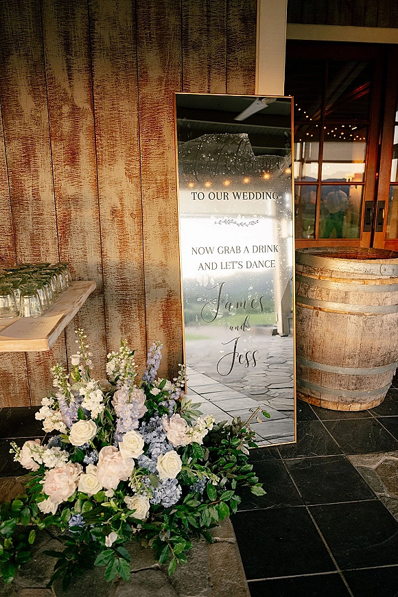 Wedding welcome sign on a gold-framed mirror with calligraphy, white roses and blue flowers, on a rustic wood wall with string lights