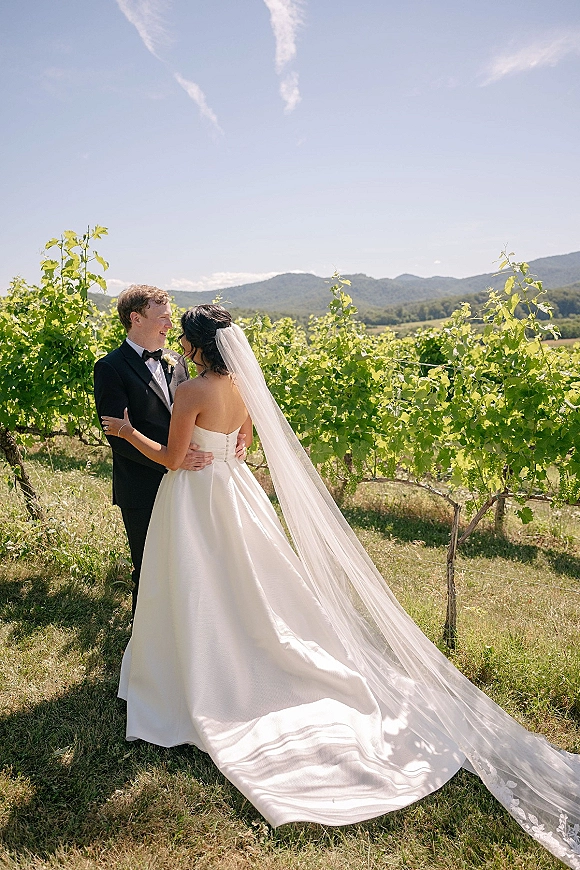 Couple portrait of bride and groom laughing in a vineyard wedding portrait, her long veil trailing in the breeze with mountains behind