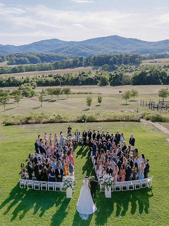Outdoor wedding ceremony with bride walking down the aisle between white chairs and floral pedestals on a green lawn with hills and mountains beyond