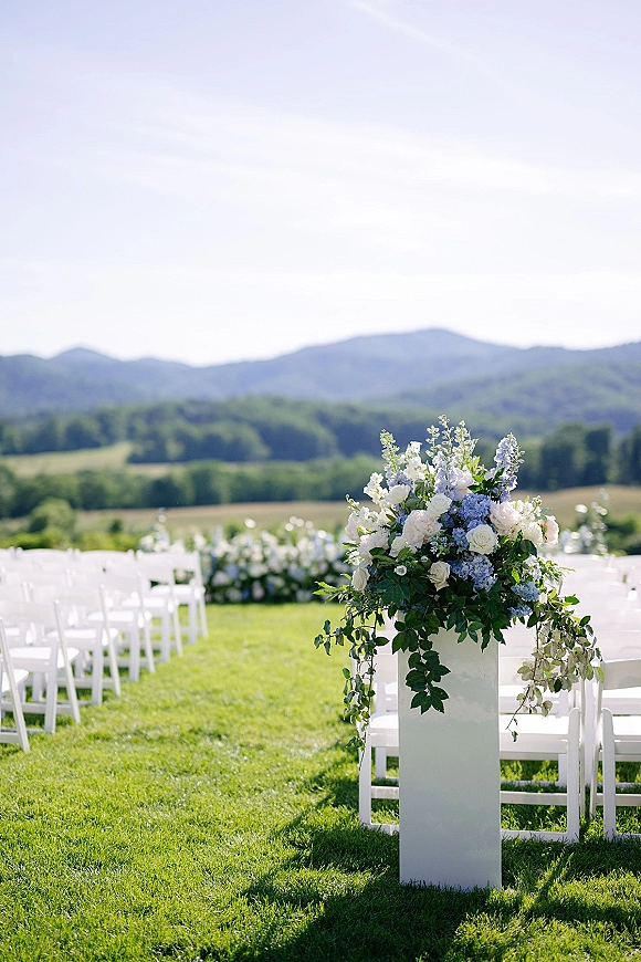 Ceremony aisle decor with white folding chairs and blue-and-white floral arrangements on a lawn, set against mountains and rolling hills
