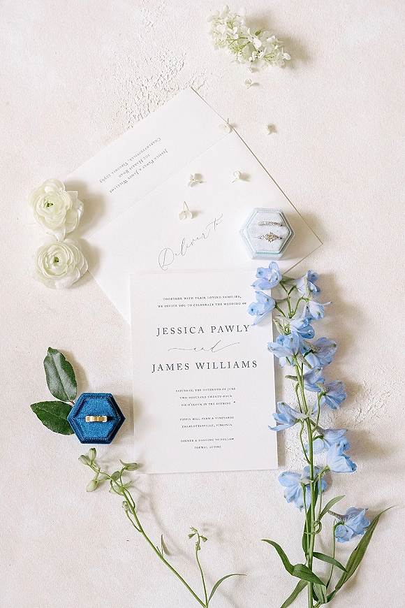 Wedding invitation suite flat lay with calligraphy cards, RSVP, envelope, and rings beside blue delphinium on a white textured surface