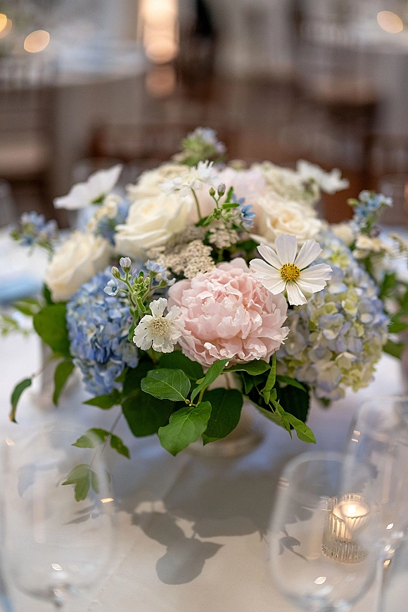 Wedding centerpiece with white roses, blue hydrangea and pink peony in a low vase on a white tablecloth with votive candle and bokeh lights