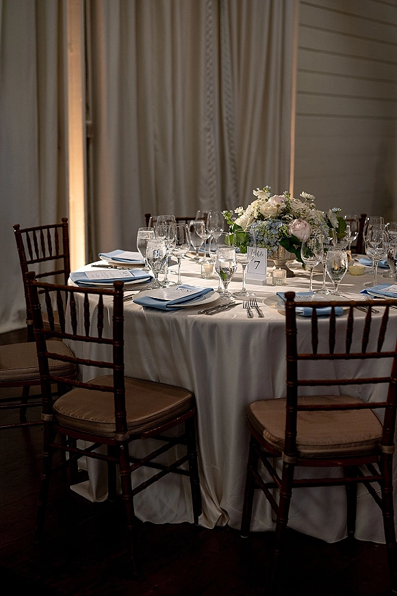 Reception tablescape with a round wedding table in white linen, rose and hydrangea centerpiece, votive candles, and chiavari chairs by draped curtains