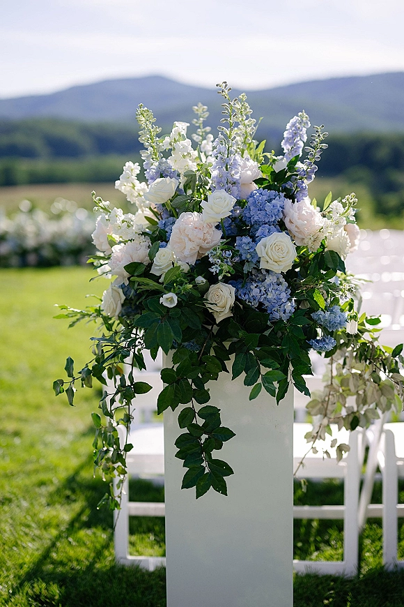 Ceremony floral arrangement on a white pedestal with roses, blush peonies, blue hydrangeas and greenery beside outdoor chairs and mountains