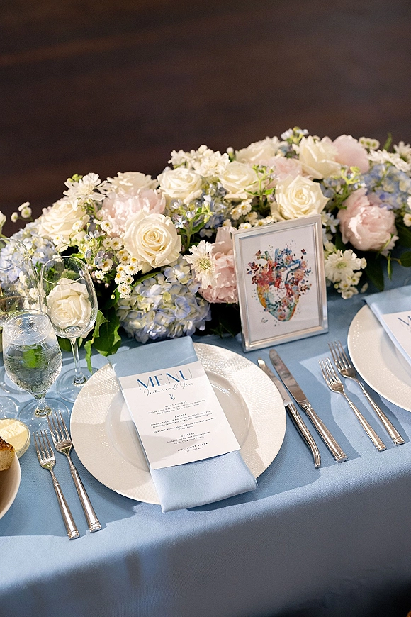 Reception tablescape with wedding table setting featuring blue linens, dusty blue napkin, menu card, framed photo, and rose hydrangea florals against a dark wall