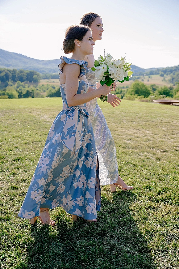 Bridesmaid walking in a blue floral dress carrying a white rose and greenery bouquet across a grassy field with distant mountains behind