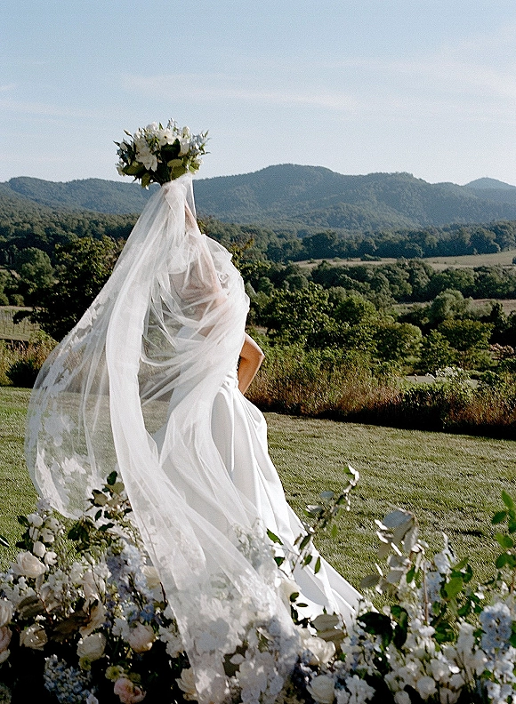 Bridal portrait of a bride holding a white and green bouquet, her long veil blowing over a strapless dress in a mountain meadow