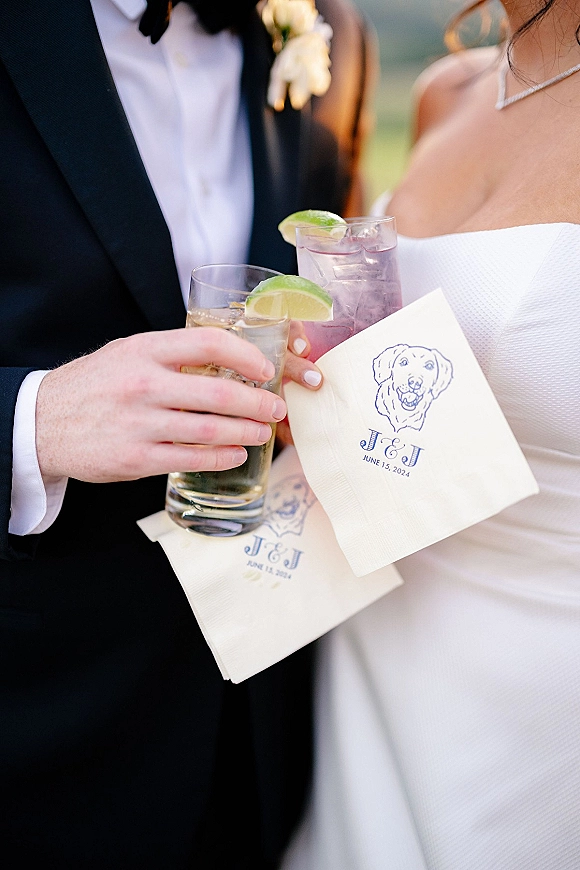 Wedding cocktail toast as bride and groom clink glasses with lime wedges, monogrammed napkins, tuxedo boutonniere, and greenery backdrop