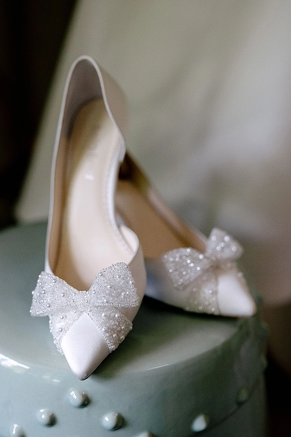 Bridal shoes with pearl bow heels and beaded embellishments, pointed toe pumps perched atop a blue frosted wedding cake indoors