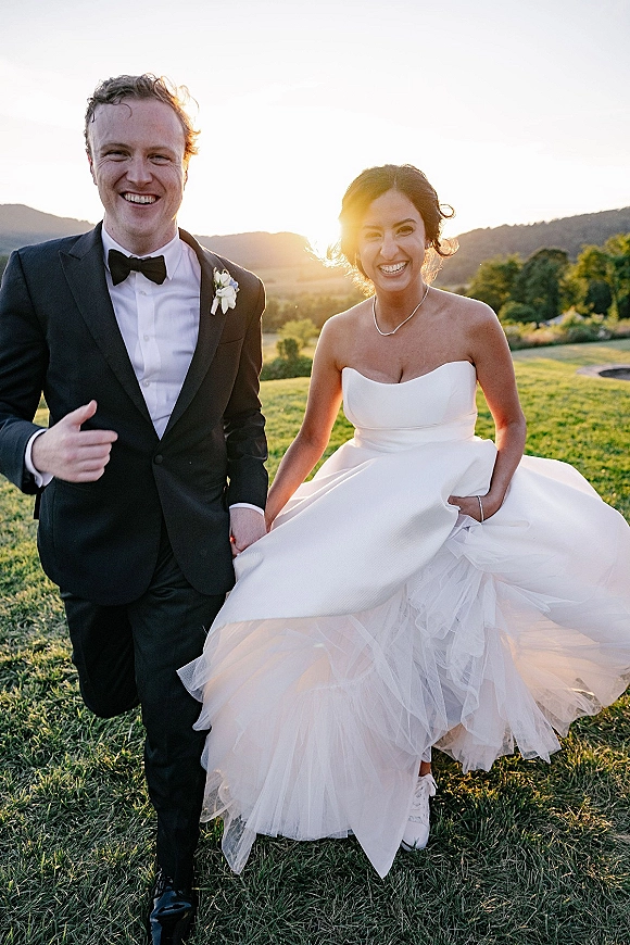 Couple portrait outdoors of bride and groom laughing at sunset, she lifts her tulle skirt beside him in tuxedo on grassy hills
