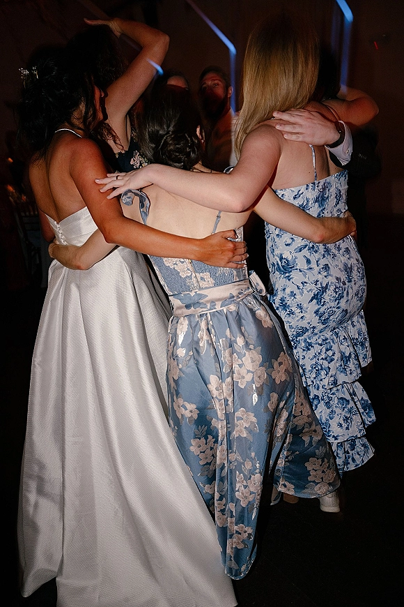 Wedding dance floor reception dance party as the bride in a strapless wedding dress is hugged by friends under party lighting in a dim room