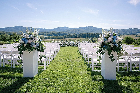 Outdoor ceremony setup with white folding chairs and blue and white floral pedestal arrangements along an aisle on a lawn with mountain views