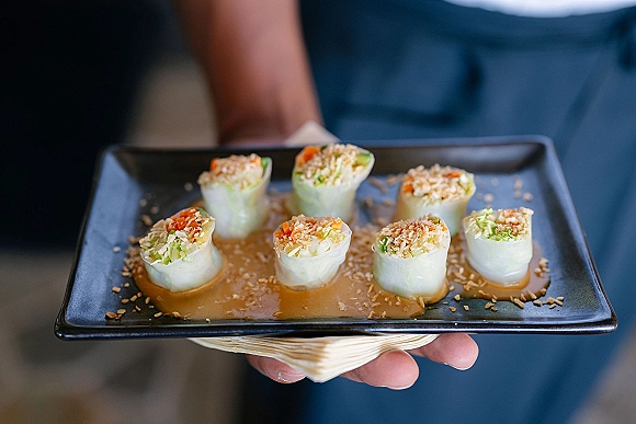 Wedding appetizers on a black serving tray, fresh spring rolls with dipping sauce and toasted coconut flakes held by a server indoors