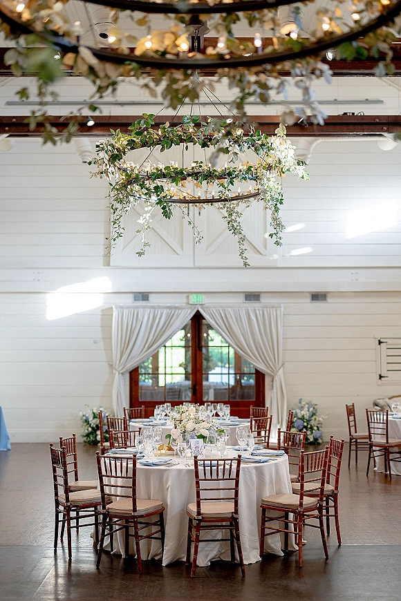 Reception tablescape with a round wedding reception table set in white linens, floral and greenery centerpiece, candles, and string lights overhead