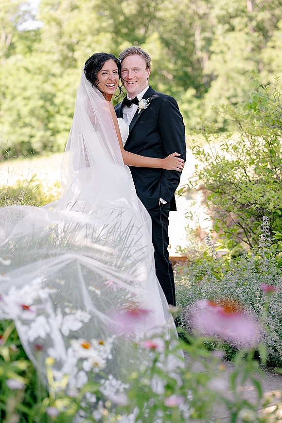 Couple portrait of bride in a strapless gown with long veil and groom in tuxedo hugging on a garden path with wildflowers