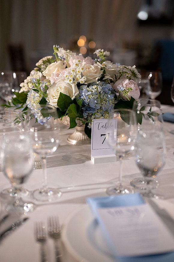 Reception tablescape with a wedding table centerpiece of white roses and blue hydrangeas in a compote vase, lit by warm bistro lights