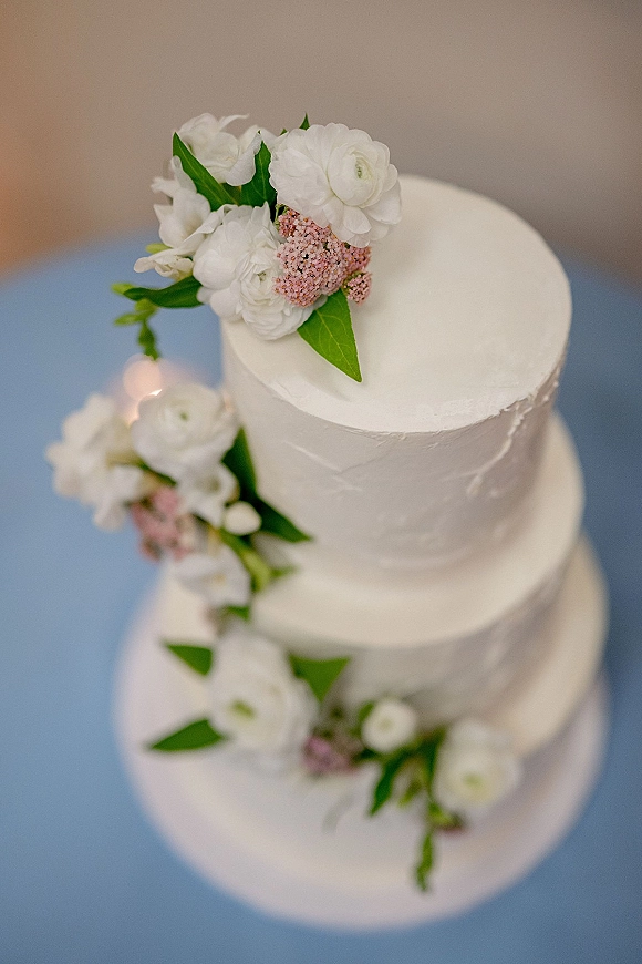 Wedding cake two tier wedding cake with white buttercream frosting, white and pink flowers with green leaves on a blue surface, candlelight bokeh behind