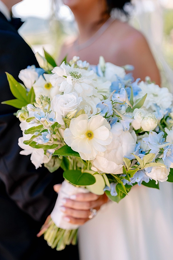 Bridal bouquet of white and blue wedding bouquet flowers with greenery and ribbon-wrapped handle, held by bride in strapless dress outdoors