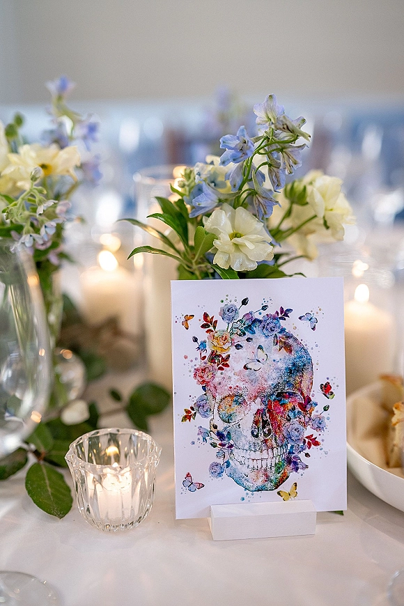 Wedding table decor with a wedding place card featuring watercolor skull art, blue and white flowers, greenery, and candlelit glassware on a reception table