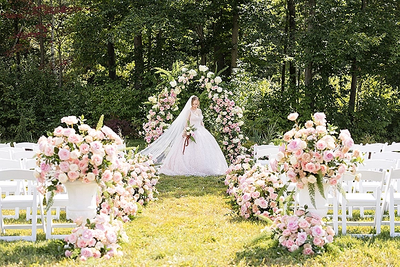 Outdoor ceremony setup with a floral arch of pink roses and greenery, white chairs and aisle pedestals on a sunlit garden lawn