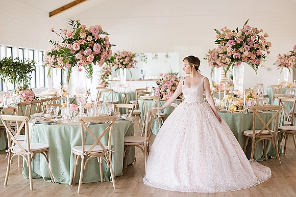 Bride portrait in a ball gown wedding dress with floral appliqué, standing by mint linen tables with blush rose centerpieces in a bright reception room