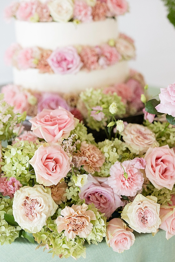 Wedding cake with floral wedding cake accents of pink roses and hydrangeas cascading over white tiers on a stand against a white backdrop